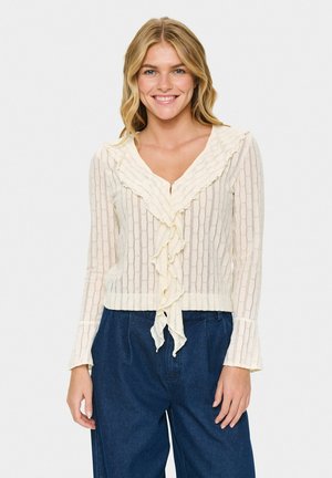Giovane donna sorridente, che indossa una camicetta crema con volant e strisce verticali e pantaloni larghi blu scuro, su uno sfondo chiaro e semplice.