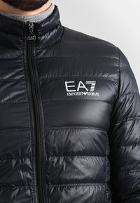 Zwarte pufferjas met horizontale stiksels, hoge kraag, volledige ritssluiting en logo aan de voorkant links. Het materiaal lijkt glanzend en glad.