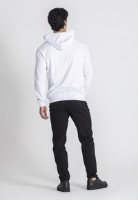 Sudadera blanca con capucha, puños y dobladillo acanalados. Llevada con jeans negros y zapatos negros. La vista es desde la parte de atrás contra un fondo simple.