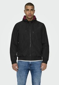 Zwarte windjack met een kastanjebruine voering in de capuchon, heeft een volledige rits, elastische tailleband en een trekkoord voor een verstelbare pasvorm.