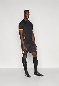 Czarna sportowa koszulka z złotymi detalami, w połączeniu z czarnymi szortami i długimi czarnymi skarpetkami. Na koszulce, skarpetkach i korkach znajduje się logo PUMA.