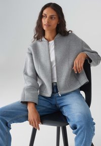 Felpa grigia cropped con colletto alto, chiusura con zip e dettagli di cucitura discreti, abbinata a jeans blu. Tessuto morbido, vestibilità rilassata.