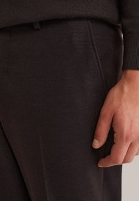 Nahaufnahme einer Hand, die neben schwarzen strukturierten Hosen liegt, wobei ein passender schwarzer Pulloverärmel sichtbar ist.