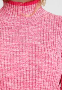 Rosa ribbat tröja med hög krage som har ett strukturerat stickmönster och en blandning av ljusare och mörkare nyanser av rosa.