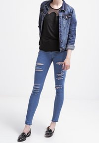 Chaqueta de mezclilla, blusa negra con detalles de malla y jeans ajustados azules desgastados con múltiples rasgaduras; combinados con mocasines negros.