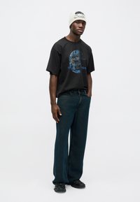 Svart oversize t-shirt med blått grafiskt tryck, kombinerad med mörkblå vida jeans. Modellen bär en ljus mössa och sneakers.