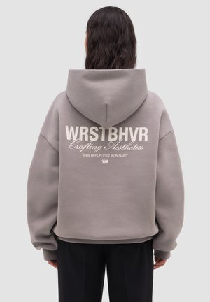 Persona con cabello largo y oscuro, vistiendo una sudadera gris de gran tamaño con el texto "WRSTBHVR Crafting Aesthetics" en la espalda, de pie frente a un fondo liso.