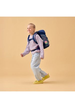 Young girl wearing a purple sweater, light jeans, yellow shoes, and a navy blue backpack, walking with a joyful expression against a beige background.