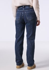 Jeans de mezclilla azul oscuro con corte recto, que cuentan con dos bolsillos traseros y detalles de costura sutiles. Combinados con un suéter crema y calzado beige.