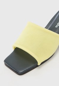 Sárga textil csúszó szandál széles pánttal és négyzet alakú orral. A talpa fekete és textúrált, finom varrási részletekkel.