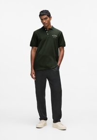 Polo vert foncé à texture douce, avec des manches courtes et un col. Pantalon noir ample et baskets beige complètent la tenue.