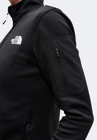Μαύρο μπουφάν North Face με φερμουάρ τσέπης στο πάνω μέρος του μανικιού και λευκό λογότυπο στο αριστερό στήθος.