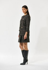 Tweedjacke mit goldenen Knöpfen, schwarz-braunem Karomuster, kombiniert mit einem gefältelten Rock und kniehohen schwarzen Stilettos.