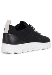 Zapatilla negra con parte superior de punto transpirable y talón de cuero negro. Presenta una suela gruesa de goma blanca y detalles de encaje plateados para mayor soporte.