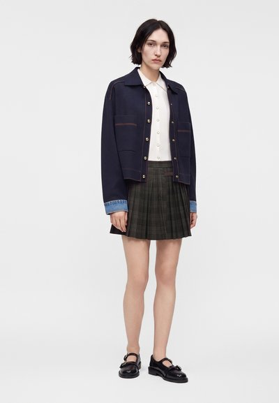 Femme portant une veste bleu marine avec des poignets en denim, une chemise blanche boutonnée, une jupe plissée foncée et des chaussures Mary Jane noires, debout devant un fond uni.