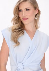 Vestito avvolgente azzurro chiaro con maniche corte, texture liscia, dettaglio in vita legato e scollatura a V, abbinato a orecchini a cerchio argentati.