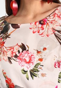 Primo piano di una blusa a fantasia floreale con fiori rosa, bianchi e rossi e foglie verdi, indossata con grandi orecchini a forma di foglia rossa.