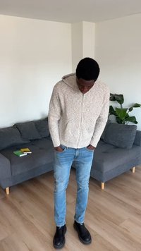 Homme en sweat à capuche beige en maille, jean bleu clair et bottes noires, debout les mains dans les poches, regardant vers le bas dans un salon avec un canapé gris.