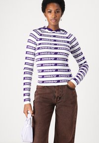 Maglietta lungo a maniche di colore viola e bianco a righe con testo "2ND DAY". Abbinata a pantaloni marroni con motivo e una piccola borsa bianca.
