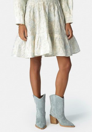 Femme portant une robe blanche brodée et des bottines en daim gris clair style western avec talons blocs en bois.