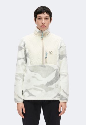 Persona joven que lleva un forro polar blanco y gris con un patrón, con un bolsillo delantero con cremallera y cuello alto, de pie frente a un fondo liso.