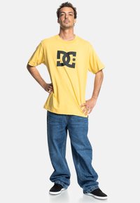 Žlté bavlnené tričko s veľkým čiernym logom DC, skombinované so voľnými modrými džínsami a čiernymi skate topánkami.