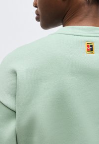 Artimas žmogaus, dėvinčio šviesiai žalią džemperį su nedideliu spalvingu Nike logotipo pleistru viršutinėje nugaros dalyje, vaizdas.