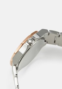 Silberne und roségoldene Uhr mit hexagonalem Gehäuse, Armband aus rostfreiem Stahl und einer glatten Textur. Sie verfügt über eine auffällige Krone.