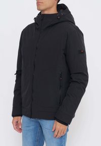 Sort vandtæt jakke med hætte, frontlynlås, sidelommer og et logopatch på ærmet. Har en glat tekstur og en tætsiddende pasform.