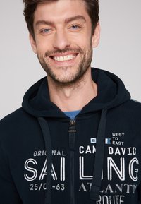 Felpa con zip di colore navy, con lettering bianco. Presenta un cappuccio con cordino, grande testo grafico e una texture morbida. Chiusure a zip visibili.