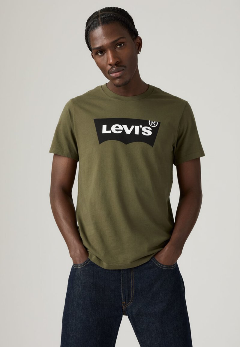 Camiseta de algodón verde oliva con un gran logo de Levi's en blanco y negro. Mangas cortas y un ajuste estándar, combinada con unos vaqueros azul oscuro.