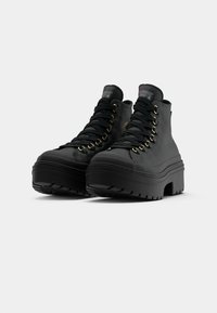 Botas altas negras con suela de plataforma gruesa, que presentan ojales dorados, cordones negros y un material superior texturizado.