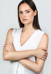Top bianco senza maniche con scollatura a V, modello con le braccia incrociate, che mostra un delicato bracciale su un polso, sfondo neutro.