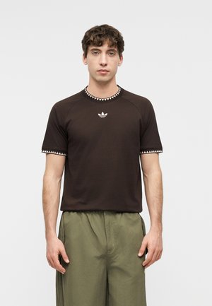 Hombre con una camiseta negra de manga corta Adidas con ribete triangular blanco y pantalones verde oliva, de pie contra un fondo claro y liso.