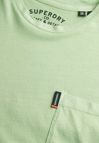 Lichtgroen katoenen T-shirt met een ronde halslijn en een borstzakje met een zwart en oranje "Superdry" label. Zachte textuur.