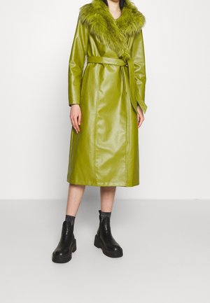 Femme portant un manteau en cuir vert mi-long avec un large col en fourrure et une ceinture, associé à des bottines noires à plateforme.