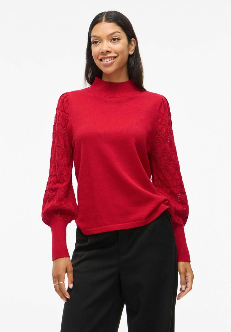 Pull rouge avec un col montant et des manches bouffantes à motifs ; le tissu du corps lisse et les manches texturées à motifs en losanges créent un contraste.