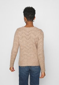 Suéter de punto en beige con textura de patrón de olas, cuello y dobladillo acanalados. Mangas largas y corte relajado. Lleva puesto unos jeans azules.