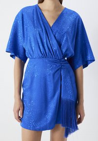 Blue wrap dress with wide sleeves, textured fabric featuring a subtle pattern, cinched waist, and a fringe detail at the hem.
