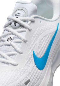 Witte sportschoen met een gestructureerde bovenkant, blauwe Nike swoosh, grijze accenten en een rond veter-systeem.