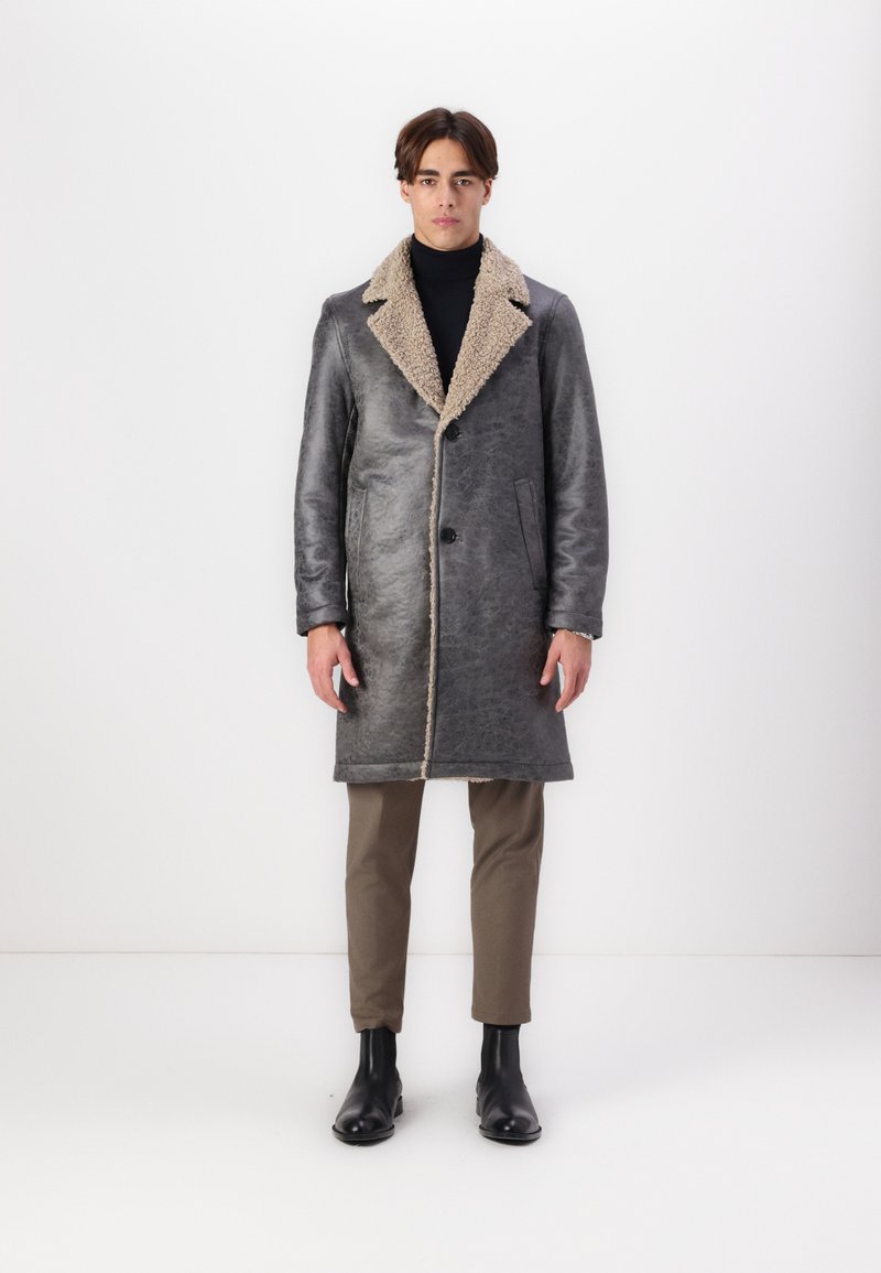 Abrigo de shearling gris con un collar beige, que presenta un cierre de botones, bolsillos laterales y un diseño largo. Llevado sobre un suéter negro de cuello alto y pantalones beige.