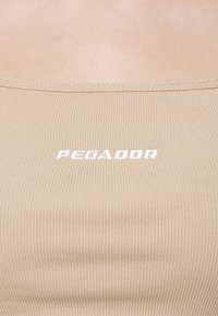 Beżowy, prążkowany materiał z bliska, z białym haftowanym tekstem "PEGADOR" umieszczonym na środku w górnej części.