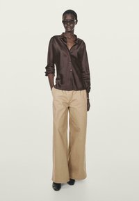 Braunes Satin-Hemd mit Knöpfen, beige weite Hosen, schwarze Schuhe und dunkle Sonnenbrille. Glatte Textur mit langen Ärmeln und leichtem Glanz.