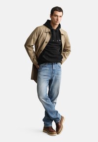 Beige trenchcoat over en sort hoodie, kombineret med lyseblå jeans og brune snørestøvler. Materialerne er bomuld, denim og læder.