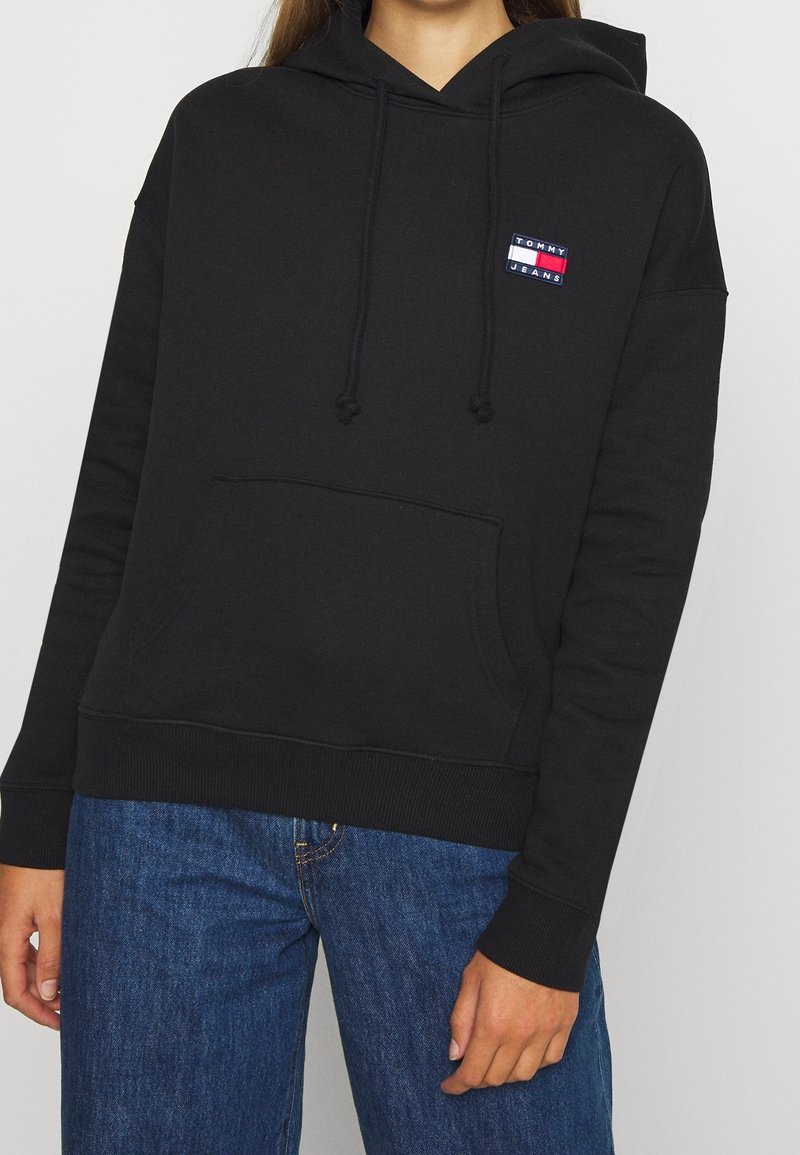 Sweatshirt à capuche noir en coton, comportant une poche frontale et un petit patch logo en rouge, blanc et bleu sur la poitrine gauche.