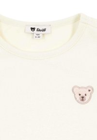 Camiseta corta de manga corta en color crema que presenta un bordado suave de la cara de un oso en el lado izquierdo del pecho. Material de algodón con un cuello redondo y costuras cosidas.
