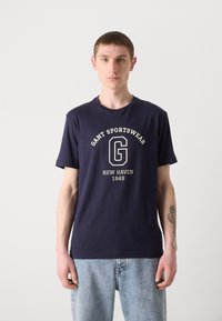 Mørkeblå bomull T-skjorte med rund hals, med en hvit logo og tekstgrafikk "GANT SPORTSWEAR NEW HAVEN 1949" foran.