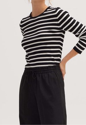 Frau trägt schwarz-weiß gestreiftes Langarmshirt mit horizontalen Streifen und lockere schwarze Hose mit Kordelzugbund, Hand auf der Hüfte ruhend.