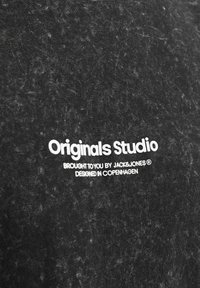 Sort stof med et struktureret mønster. Hvidt trykt logo med teksten "Originals Studio" samt yderligere tekst vedrørende brand og designplacering.