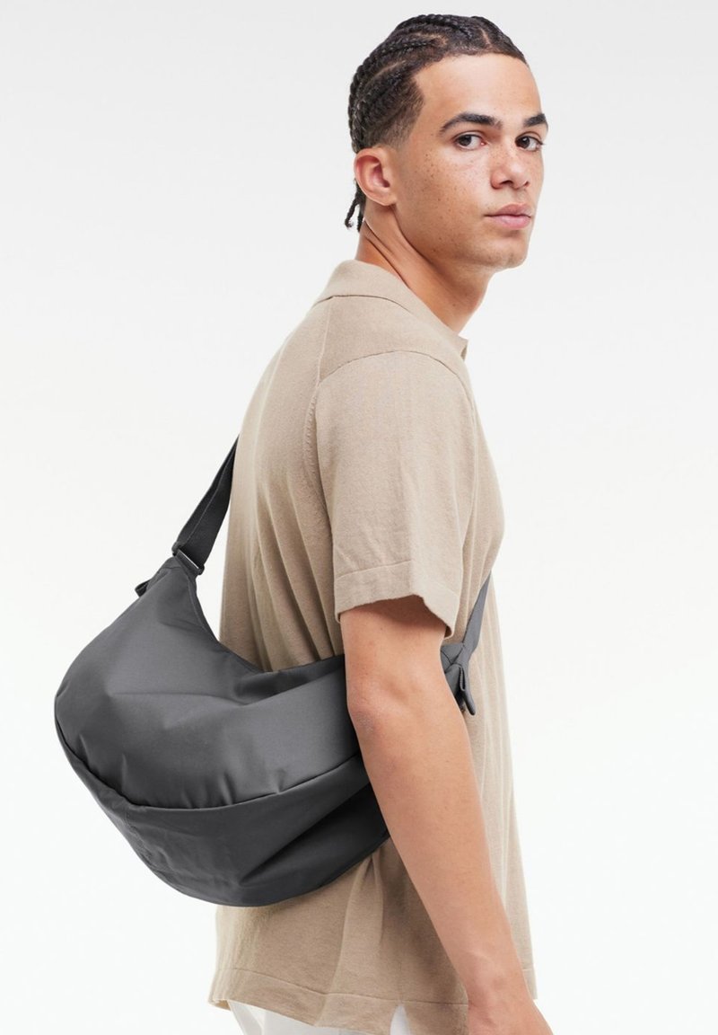 Borsa a tracolla grigia realizzata in tessuto morbido; presenta una forma curvata, una tracolla regolabile e una superficie liscia. Indossata trasversalmente per un supporto ottimale.